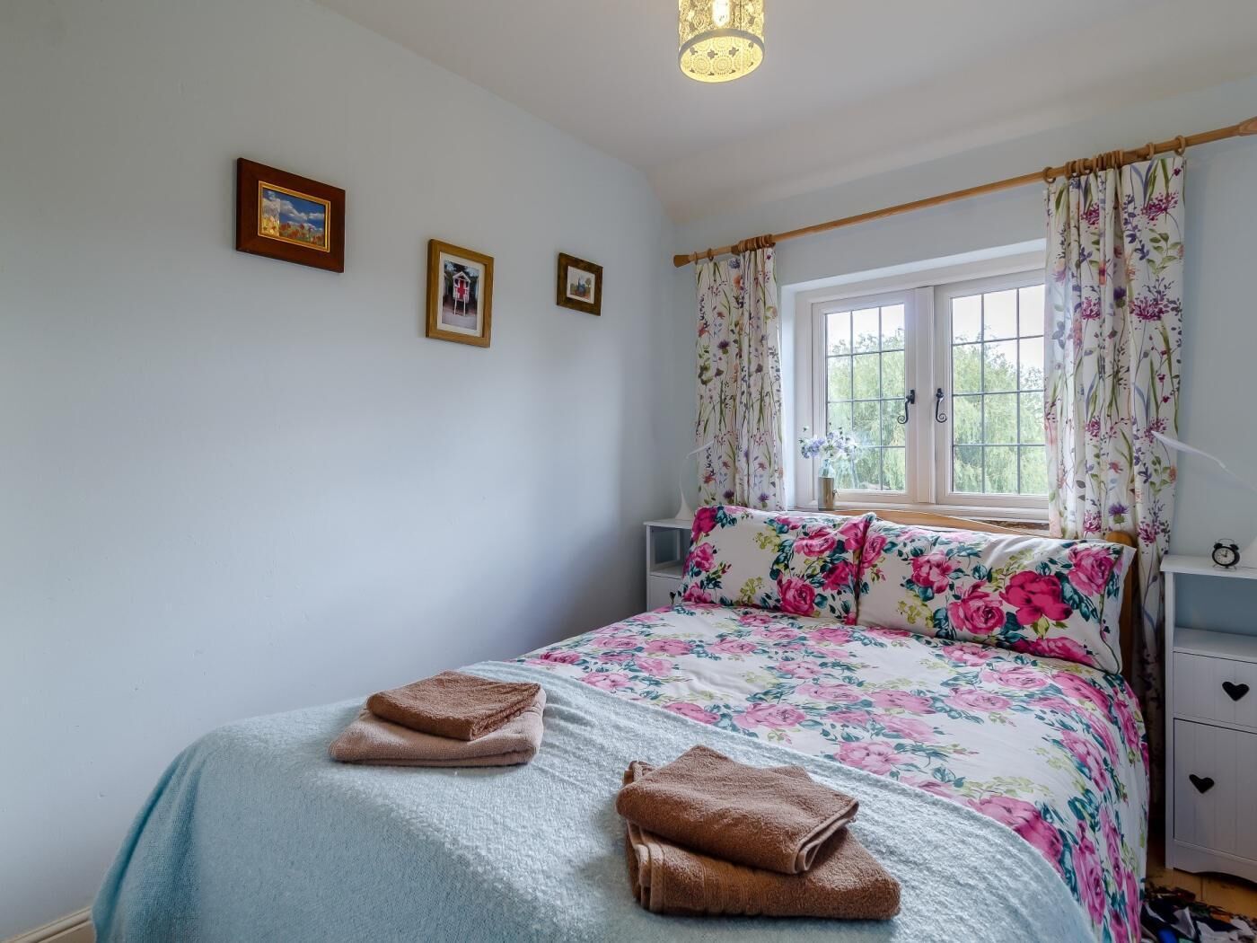A bedroom with a bed, a window with curtains, a bedside table, and a wall with three framed pictures.