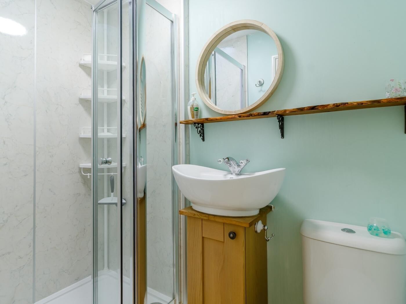 A bathroom with a glass shower enclosure, a white sink with a wooden cabinet, a round mirror, and a toilet.
