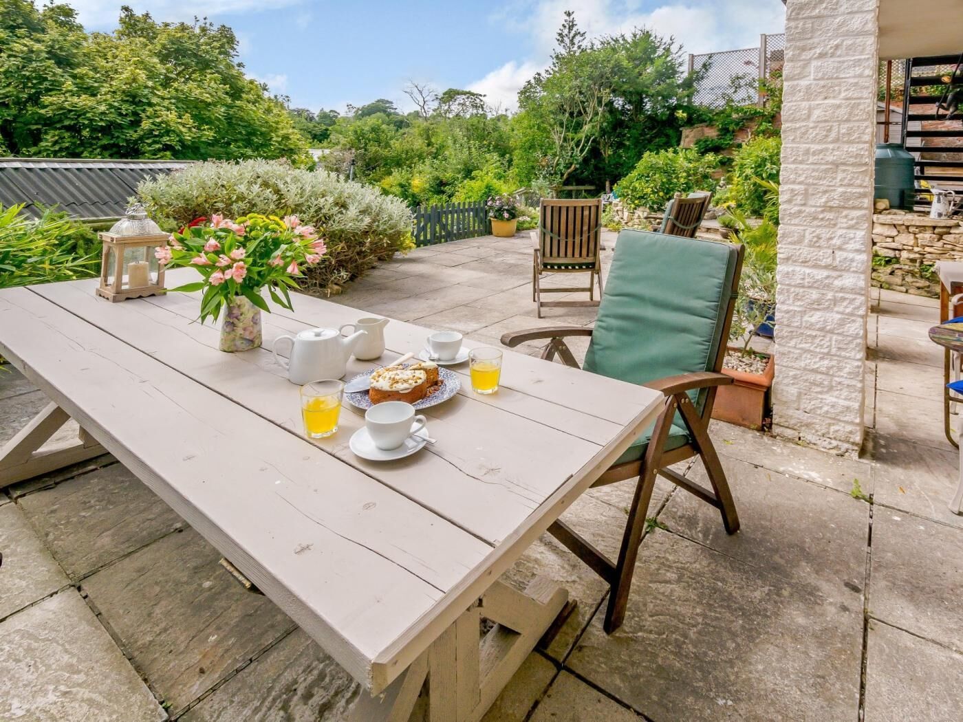 A wooden table set outdoors with a teapot, cups, a glass of tea, and a slice of cake.