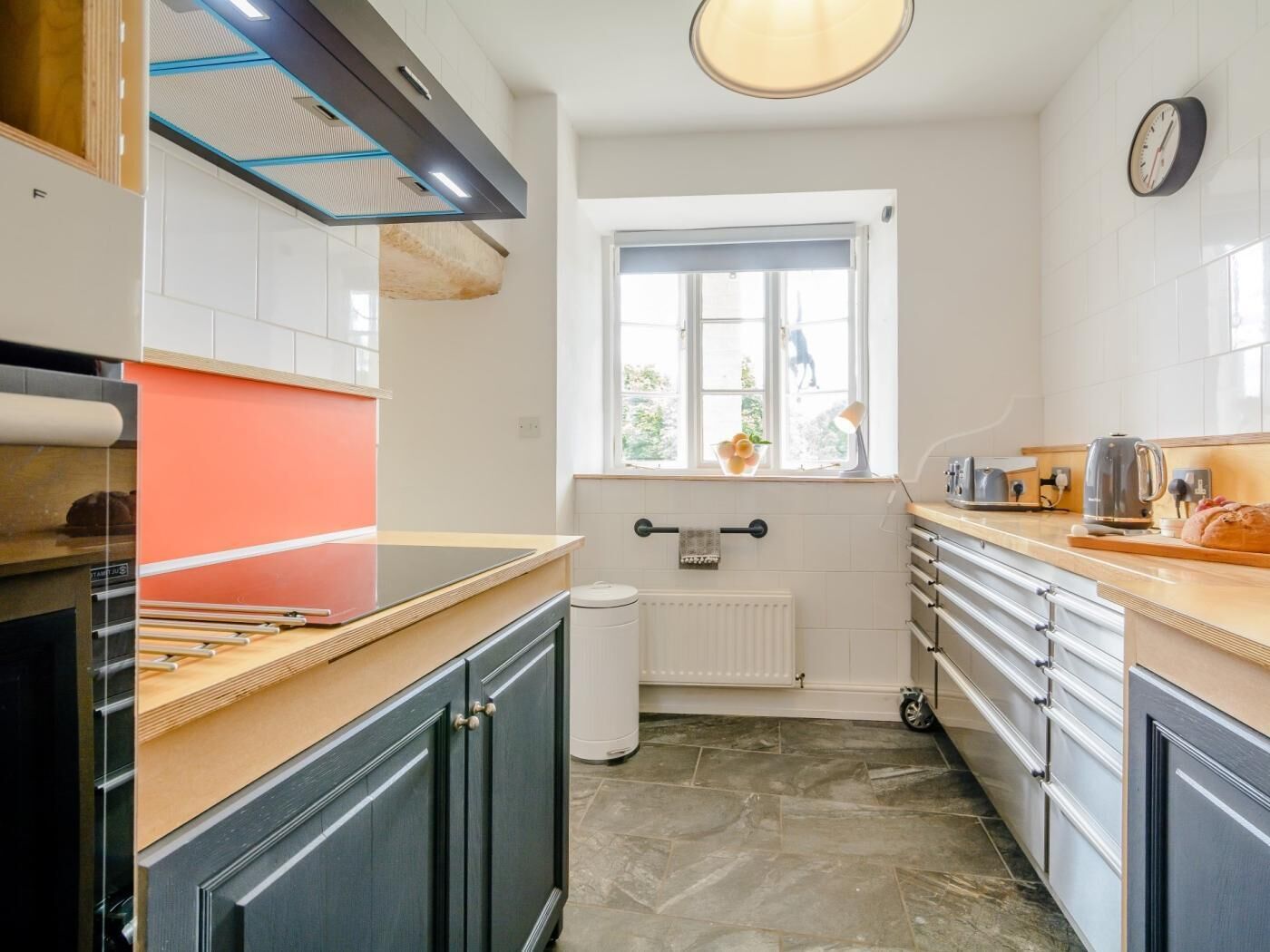 A kitchen with a window, a radiator, a clock, and a toaster.