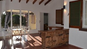 Ein Zimmer mit einem Holzkasten, einem runden Tisch mit Stühlen, einer großen Glastür, die nach draußen führt, und einem kleinen Fenster mit Rollläden.