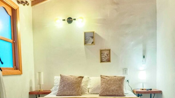 Un dormitorio con techo de madera, una cama con ropa de cama blanca, dos almohadas, una mesita de noche, una lámpara de pared y una ventana con persianas.