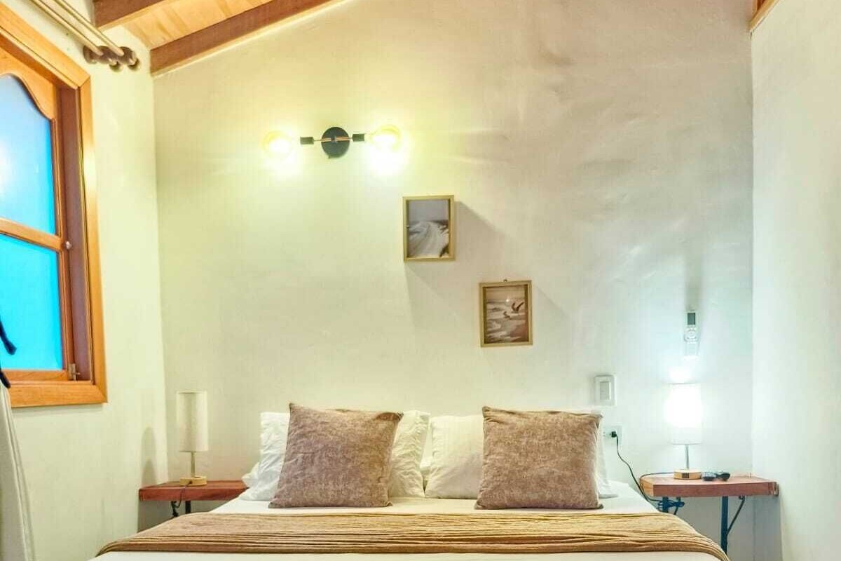 Ein Schlafzimmer mit Holzboden, einem Bett mit weißen Bettbezügen, zwei Kissen, einem Nachttisch, einer Wandleuchte und einem Fenster mit Jalousien.