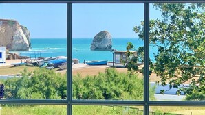Vista costera a través de una ventana: barcos, un acantilado rocoso y una playa.