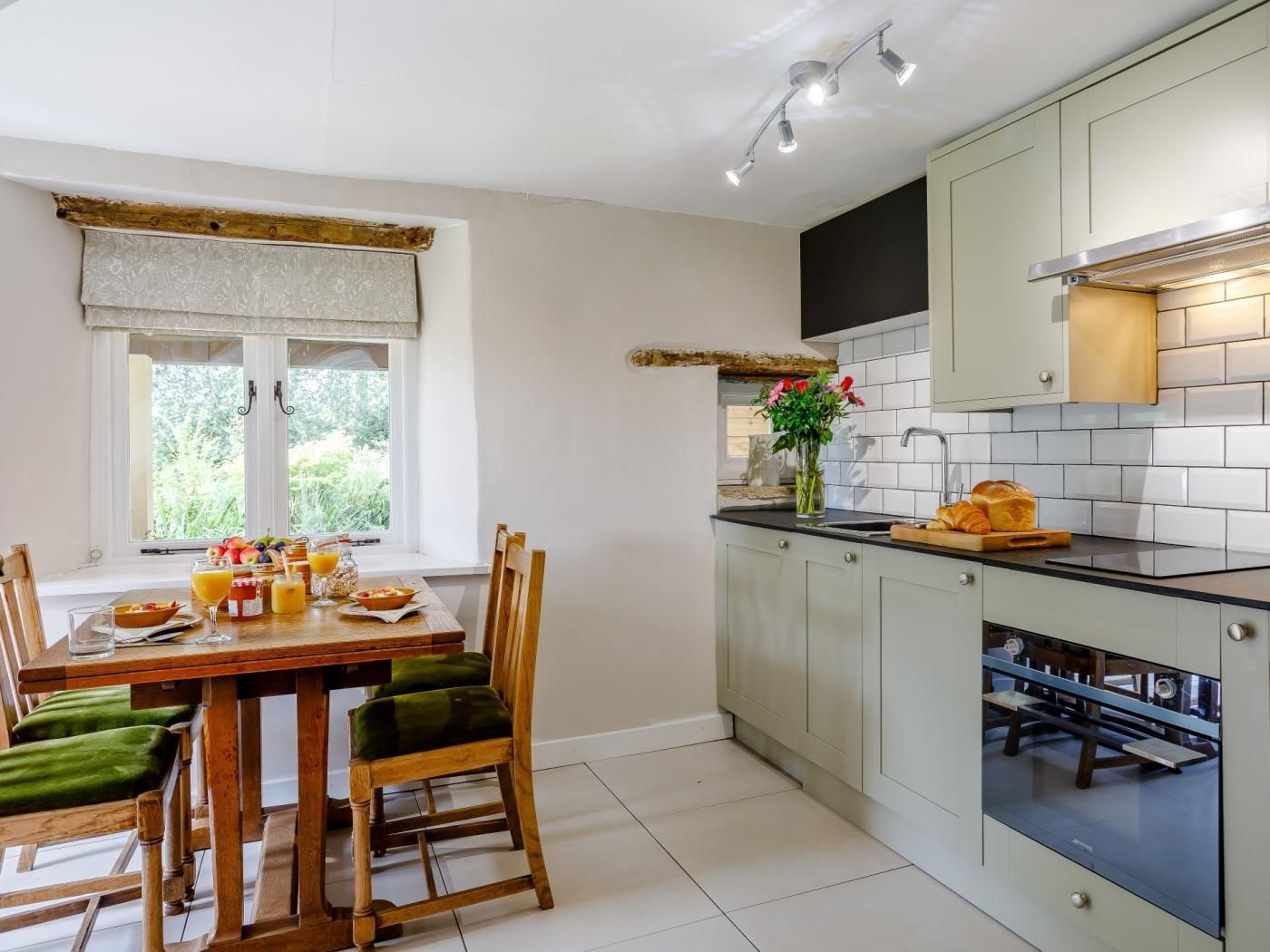 A kitchen with a dining area, featuring a window with curtains, a table set with breakfast, and a view of greenery outside.