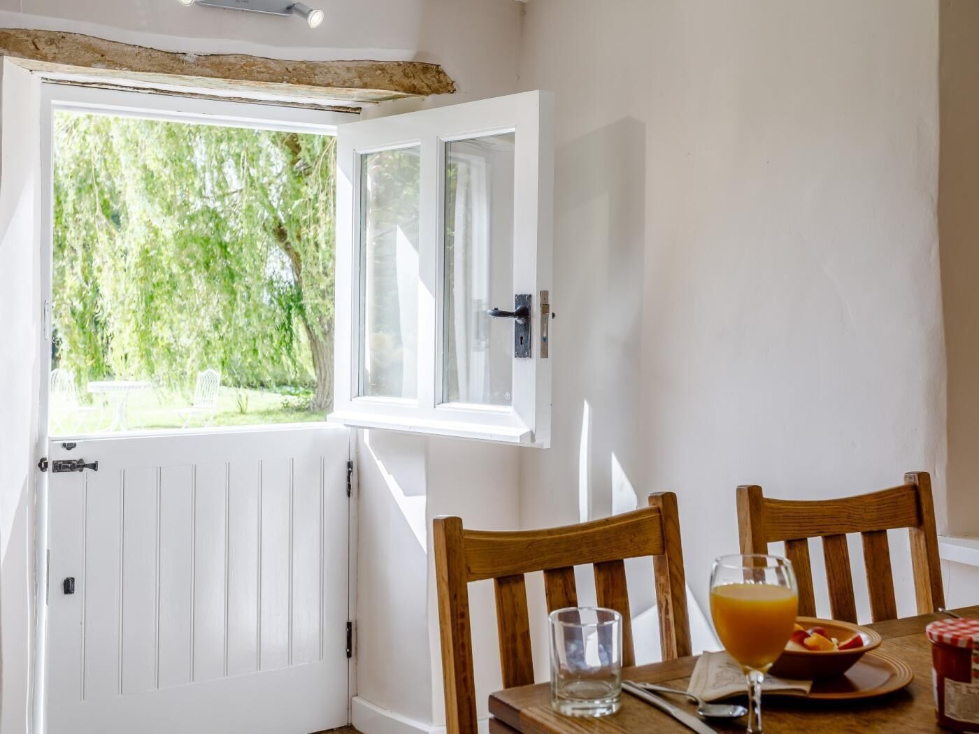 A room with a white door, a window with a view of greenery, a wooden table with a glass of orange juice, and two wooden chairs.