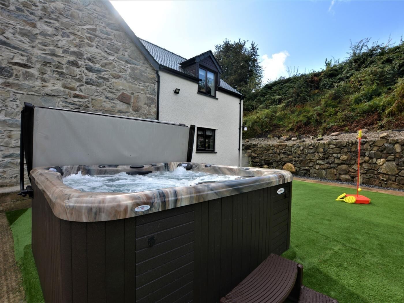 A hot tub with a stone wall backdrop, a wooden deck, and a grassy area with a golf tee.