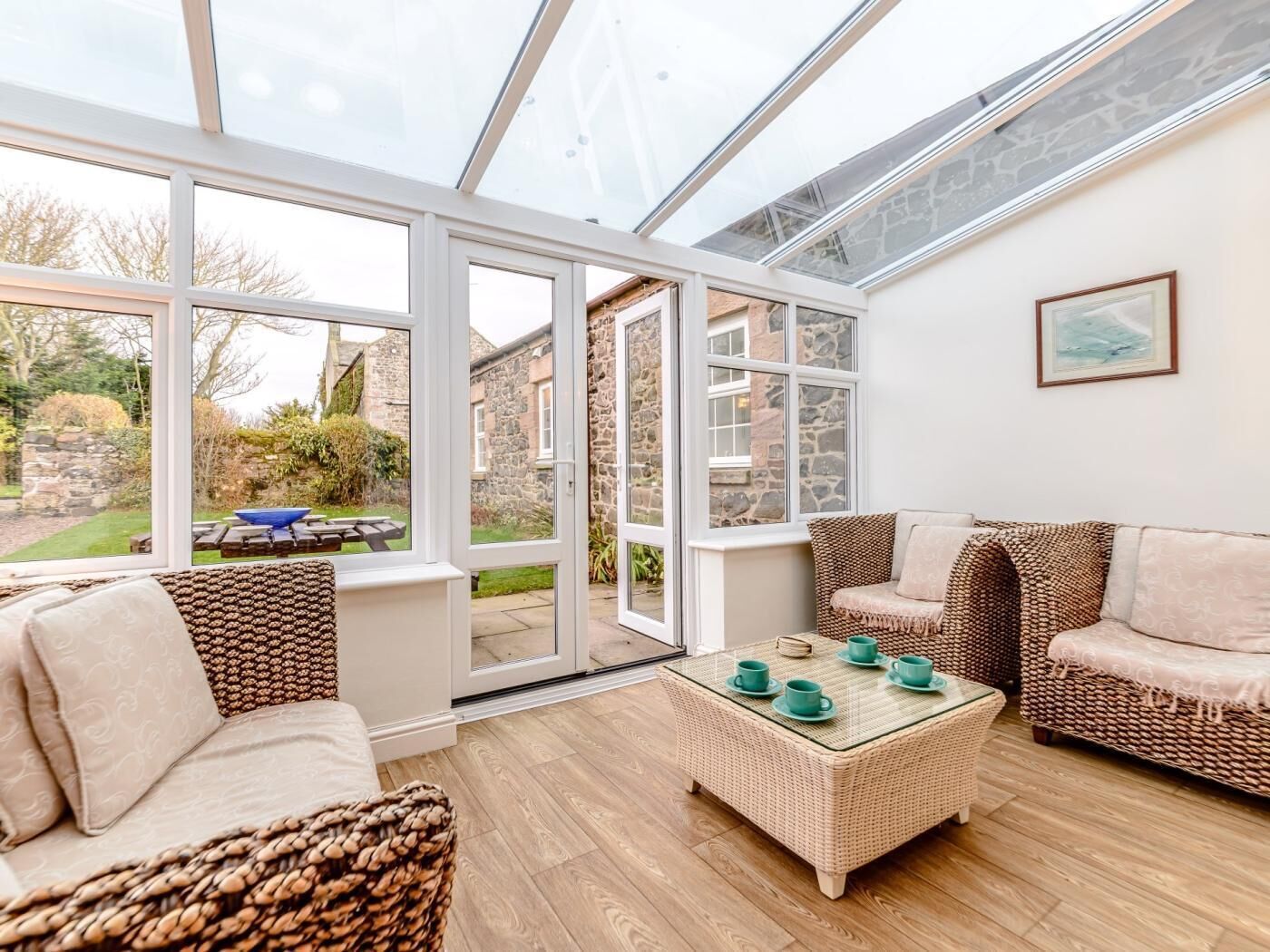 A conservatory with a glass roof, wicker furniture, and a view of a stone building.