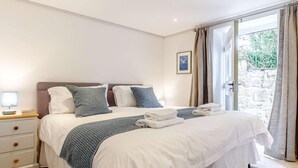 A neatly made bed with white and blue bedding, a bedside lamp, and a view of a stone wall through a window. - 2 Bed in St. Austell (oc-79594) (Coombe)