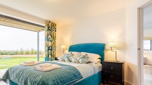 A bedroom with a bed, two bedside lamps, a nightstand, and a view of a garden. - 4 Bed in Bideford (oc-72587) (Northam)
