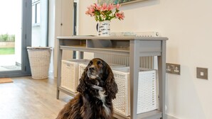 A dog sitting on a wooden floor in a room with a grey console table, wicker baskets, and a vase with flowers. - 4 Bed in Bideford (oc-72587) (Northam)