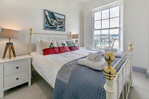 Ein ordentlich eingerichtetes Schlafzimmer mit einem Bett, Nachttischen, einem Fenster mit Blick auf die Stadt und einem Bild an der Wand.