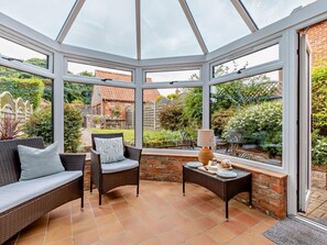 A conservatory with a glass roof, wicker furniture, a table with plates, and a view of a garden and buildings. - 3 Bed in Sheringham (oc-ncc77) (Upper Sheringham)