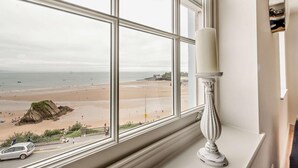 Ein Zimmer mit großem Fenster und Blick auf einen Strand, eine weiße Kerze im Fensterbrett und ein geparktes Auto vor dem Haus.