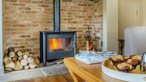 A cozy room with a brick fireplace, a wooden table with books and a bowl of cookies, and a wicker basket. - 1 Bed in Stillington (oc-g0057) (Marton-in-the-Forest)