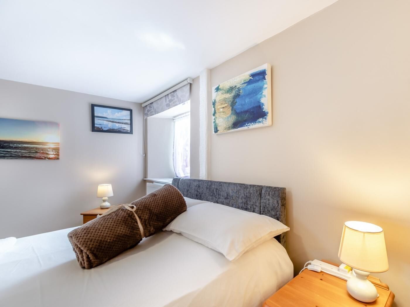 A hotel room with a bed, a nightstand, a lamp, and two paintings on the wall.