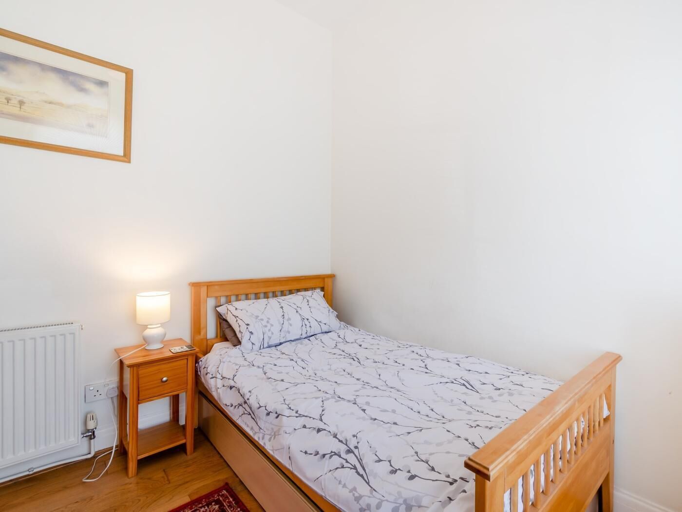Une chambre à coucher avec un lit en bois, une table de chevet avec une lampe, un radiateur et un tableau encadré au mur.