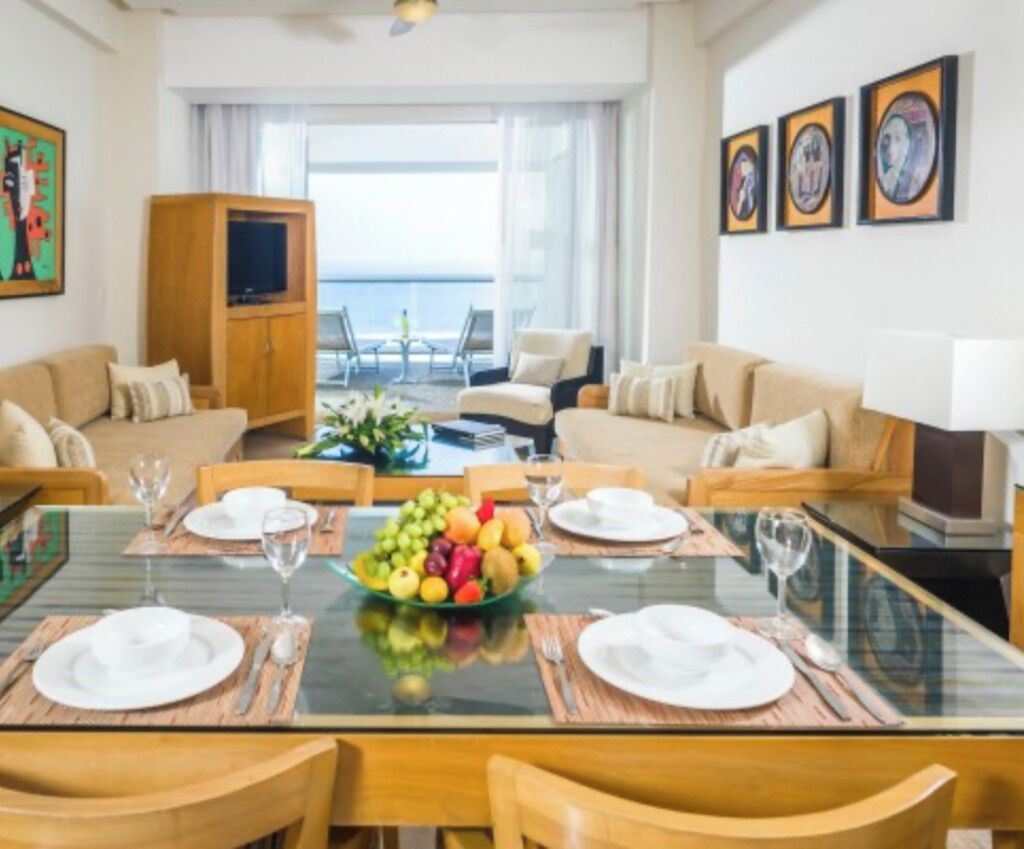 A dining area with a glass table, wooden chairs, and a fruit bowl. There is a sofa and a television in the background.