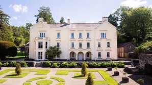 A large, cream-colored building with multiple windows and a symmetrical facade, surrounded by a well-manicured garden with geometric patterns and a stone pathway. - 4 Bed in Brecon (oc-82529) (Crai)