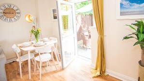 Un coin repas avec une table dressée pour quatre personnes, une horloge au mur et une vue sur l’extérieur par une porte ouverte.