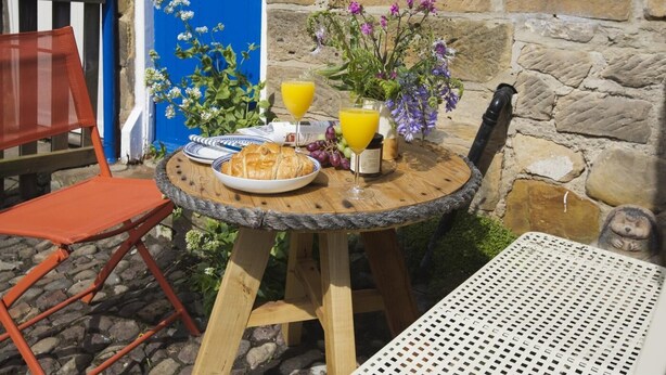 Une table ronde en bois, sur laquelle se trouvent un verre de jus d’orange, une assiette de nourriture et un vase rempli de fleurs.