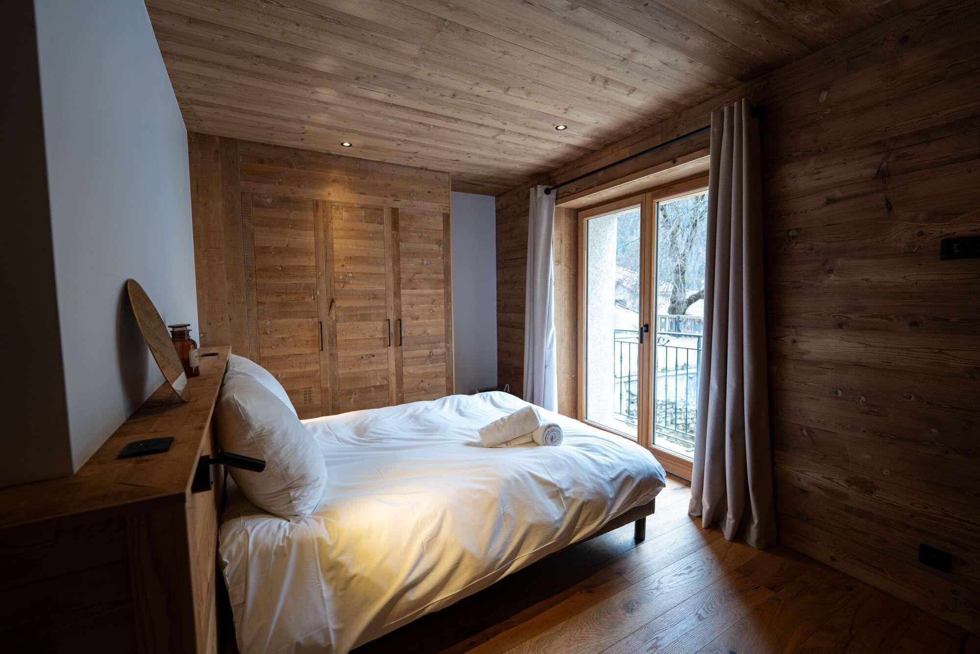 Un dormitorio con techo de madera, cama con ropa blanca, ventana con cortinas y piso de madera.