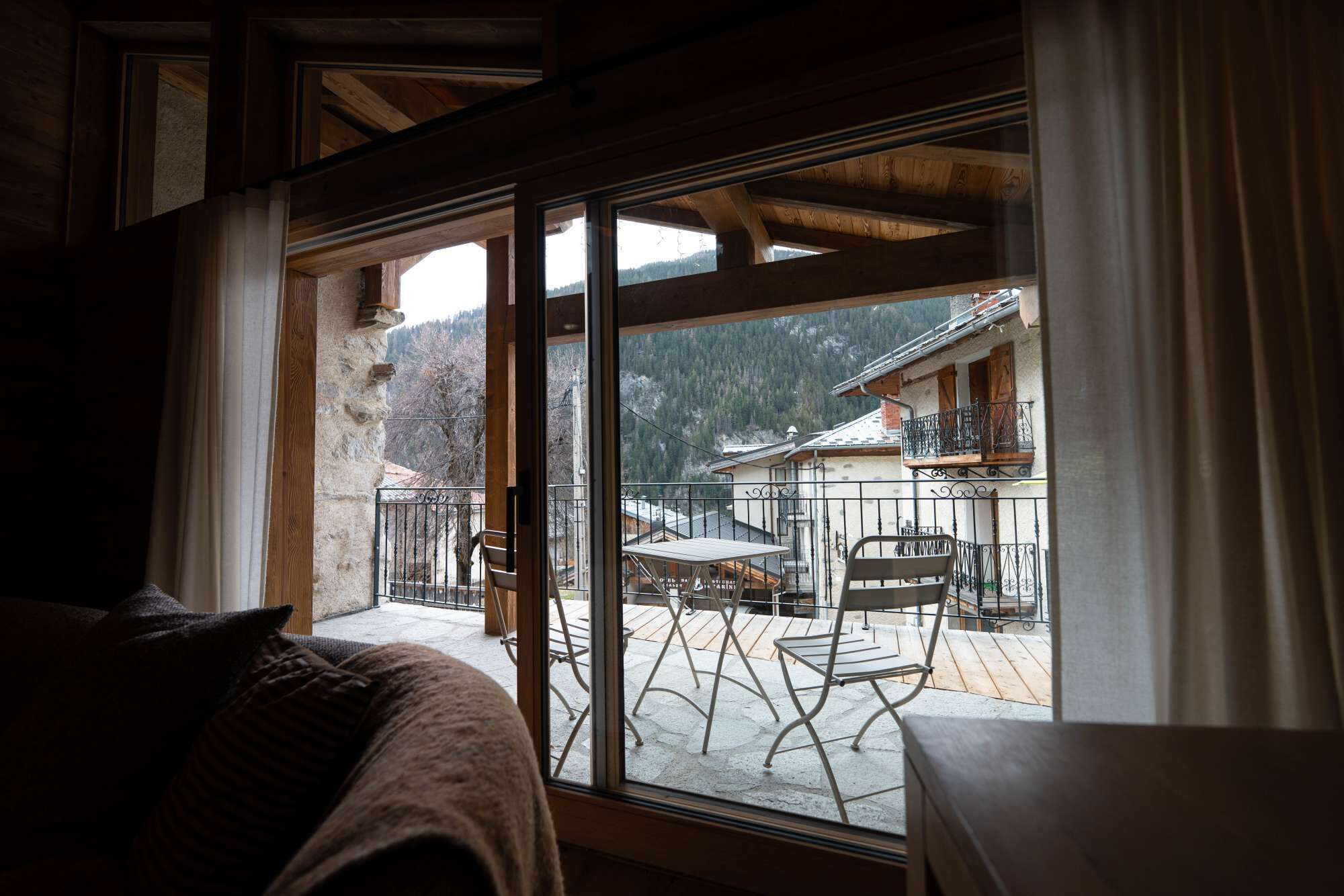 A room with a large window, wooden beams, a stone wall, and a view of a mountain and buildings.