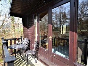 Uma cabana de madeira com porta de vidro, um pequeno alpendre com mesa e cadeiras, e vista para árvores e um carro ao fundo.