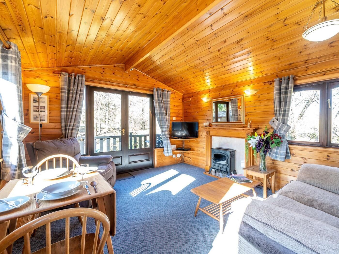 A wooden cabin interior with a sofa, dining table, TV, fireplace, and large windows.