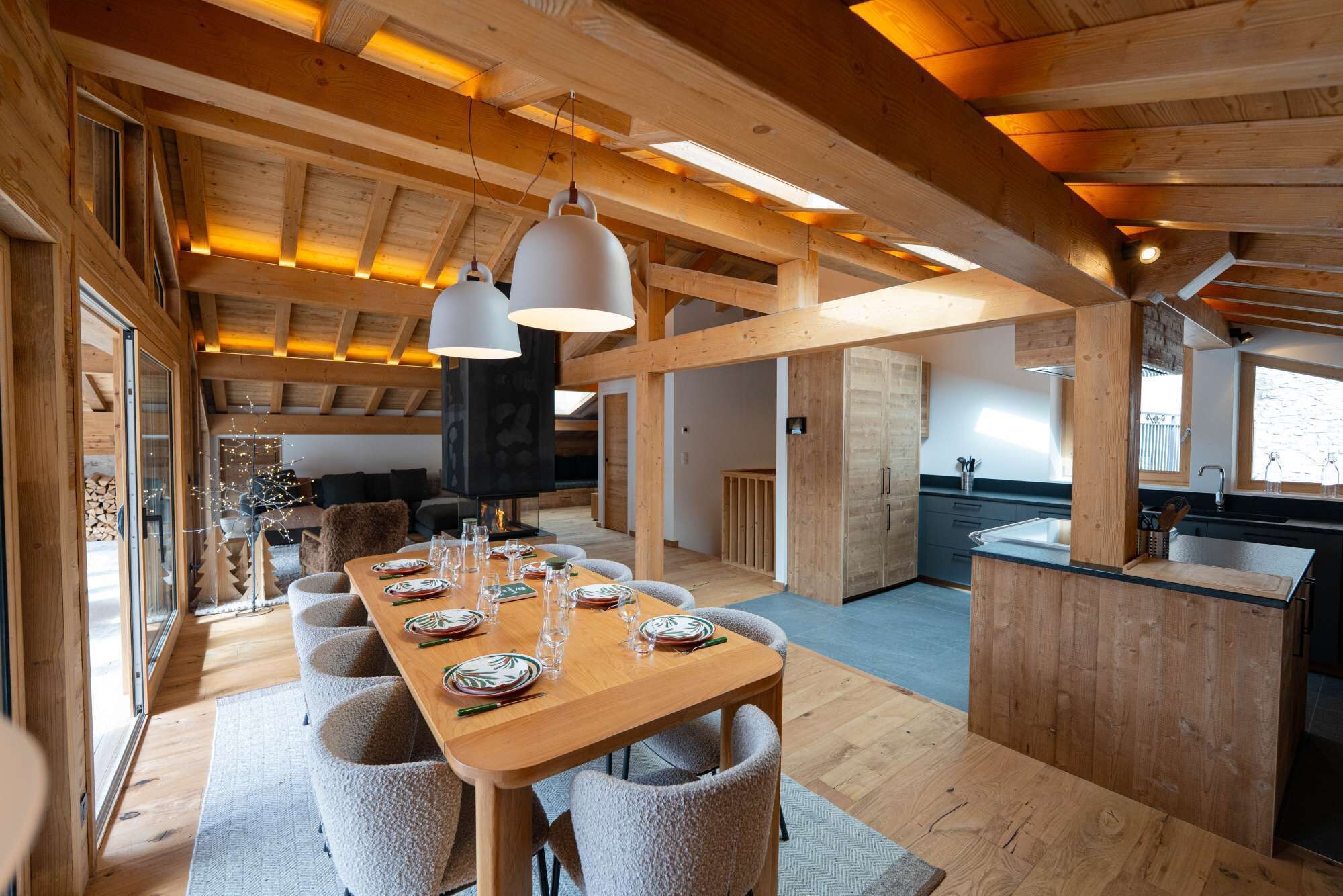 A spacious dining area with a long wooden table, multiple chairs, pendant lighting, and a kitchen in the background.