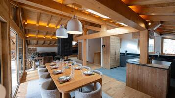 A spacious dining area with a long wooden table, multiple chairs, pendant lighting, and a kitchen in the background.