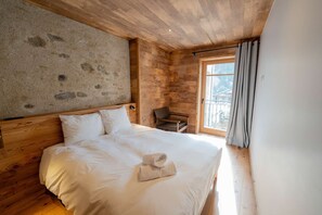 A wooden cabin bedroom with a stone wall, a bed with white linens, a chair, and a window with curtains. - Welcome to this charming, fully renovated 250-square-meter chalet right in the heart of the village. (Peisey-Nancroix)