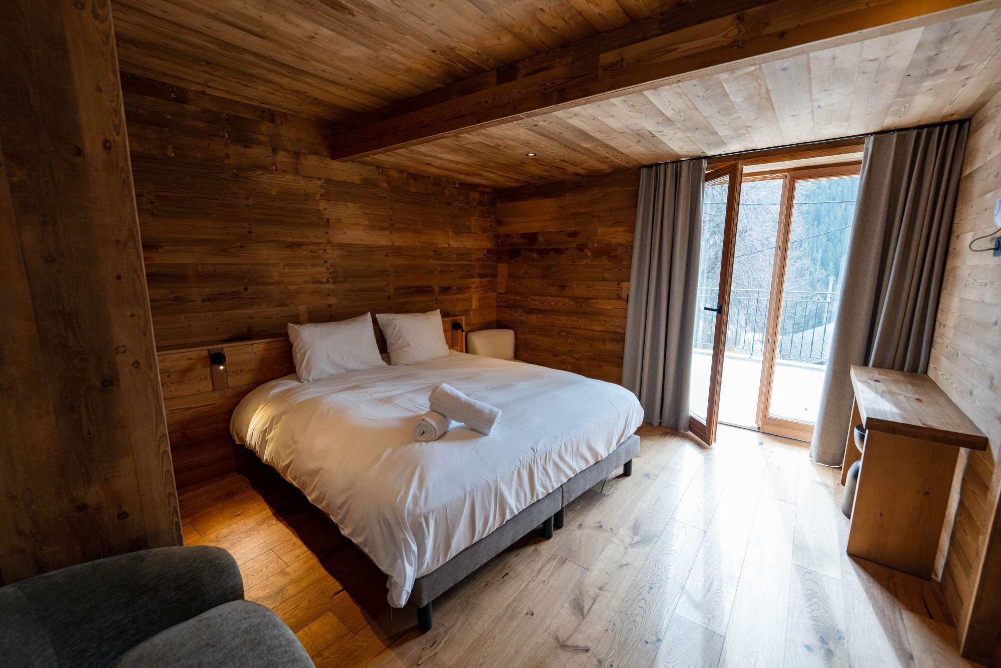 Quarto de cabana de madeira com uma cama, uma mesinha e vista através de uma porta de vidro.