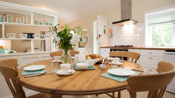 A dining area with a round table, chairs, a vase of flowers, and a kitchen in the background. - 2 Bed in Howden (oc-g0104) (Saltmarshe)