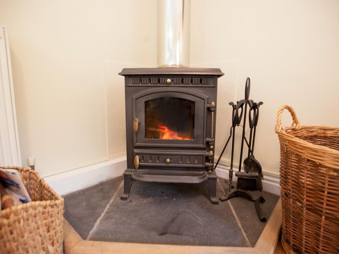 A black wood-burning stove with a fire, a wicker basket, and a metal stand.