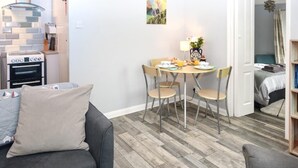 Un soggiorno moderno con un divano grigio, un tavolo da pranzo con quattro sedie, una cucina con forno e fornelli, e una camera da letto visibile attraverso una porta aperta.