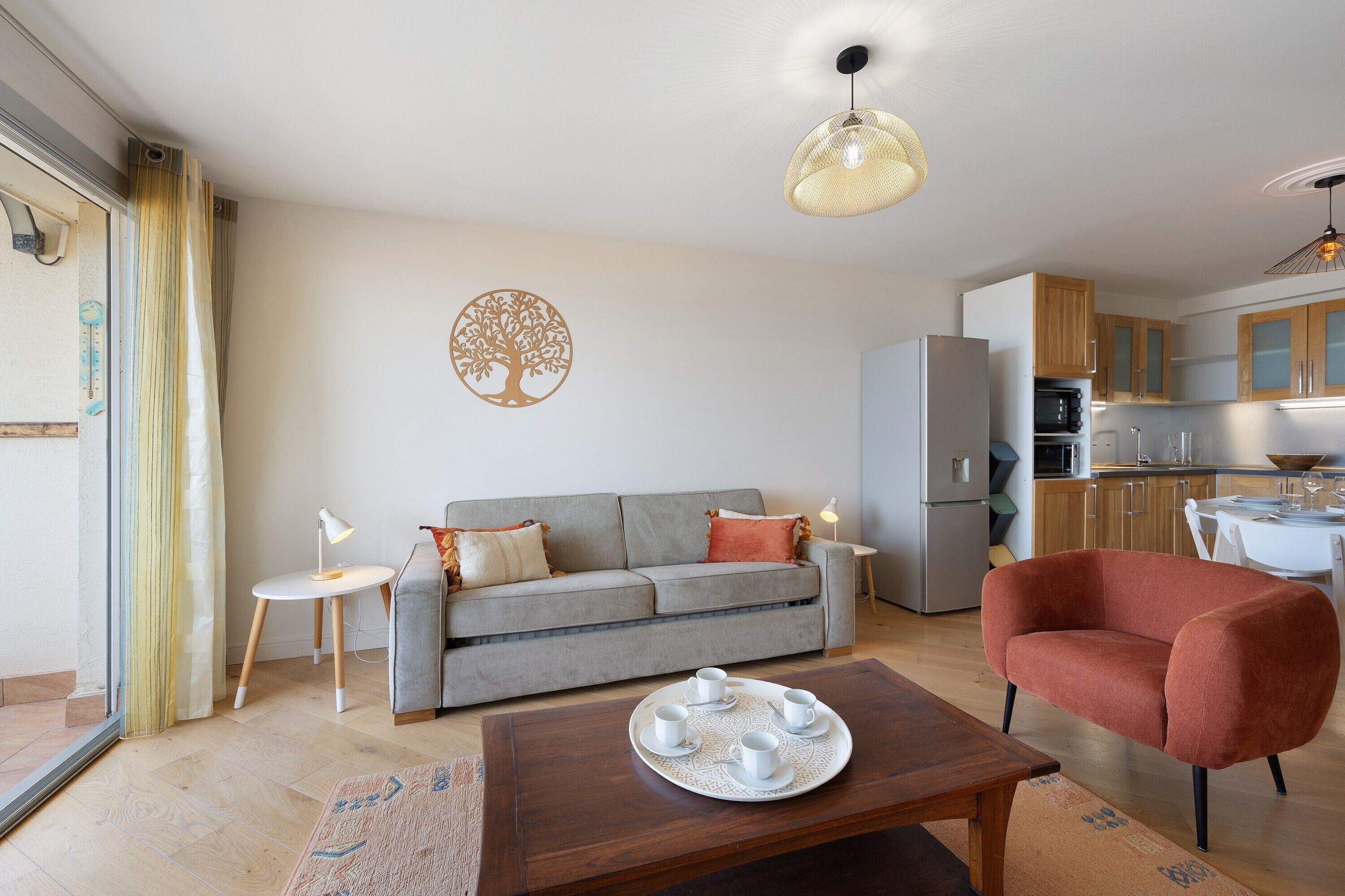 Un salon moderne avec un canapé gris, un fauteuil rouge, une table basse et une cuisine en arrière-plan.
