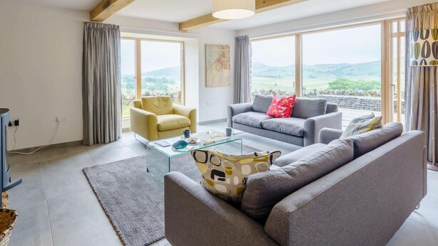 A modern living room with a sofa, armchair, and coffee table. Large windows offer a view of the countryside. - 3 Bed in Matterdale (oc-83002) (Ullswater)