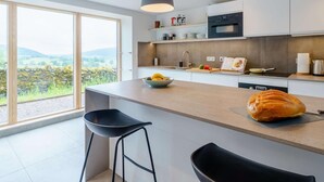 A modern kitchen with a large island, two bar stools, a view of the countryside, and a loaf of bread on the counter. - 3 Bed in Matterdale (oc-83002) (Ullswater)