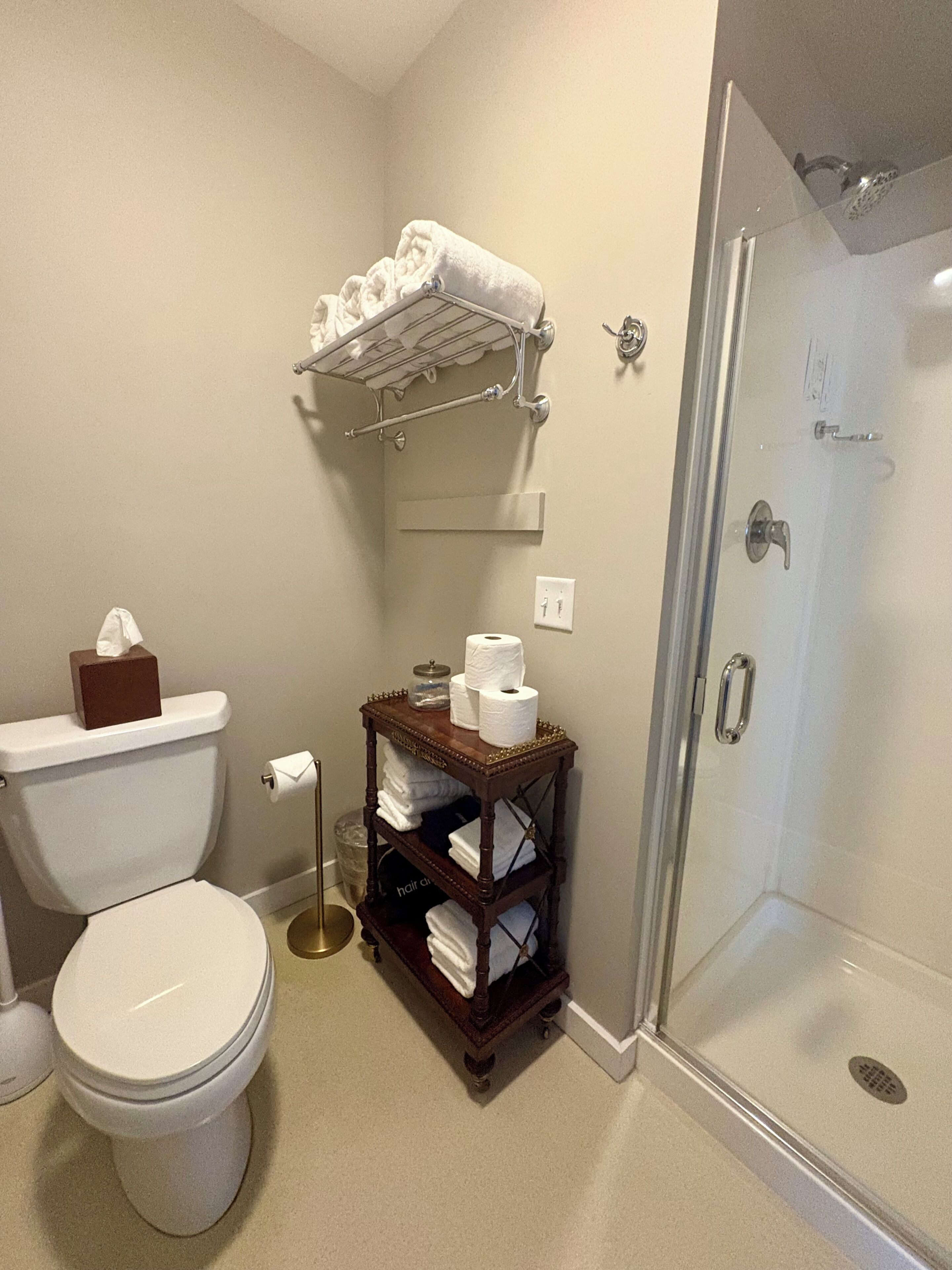 A bathroom with a toilet, a shower, and a wooden shelf with towels and toiletries.
