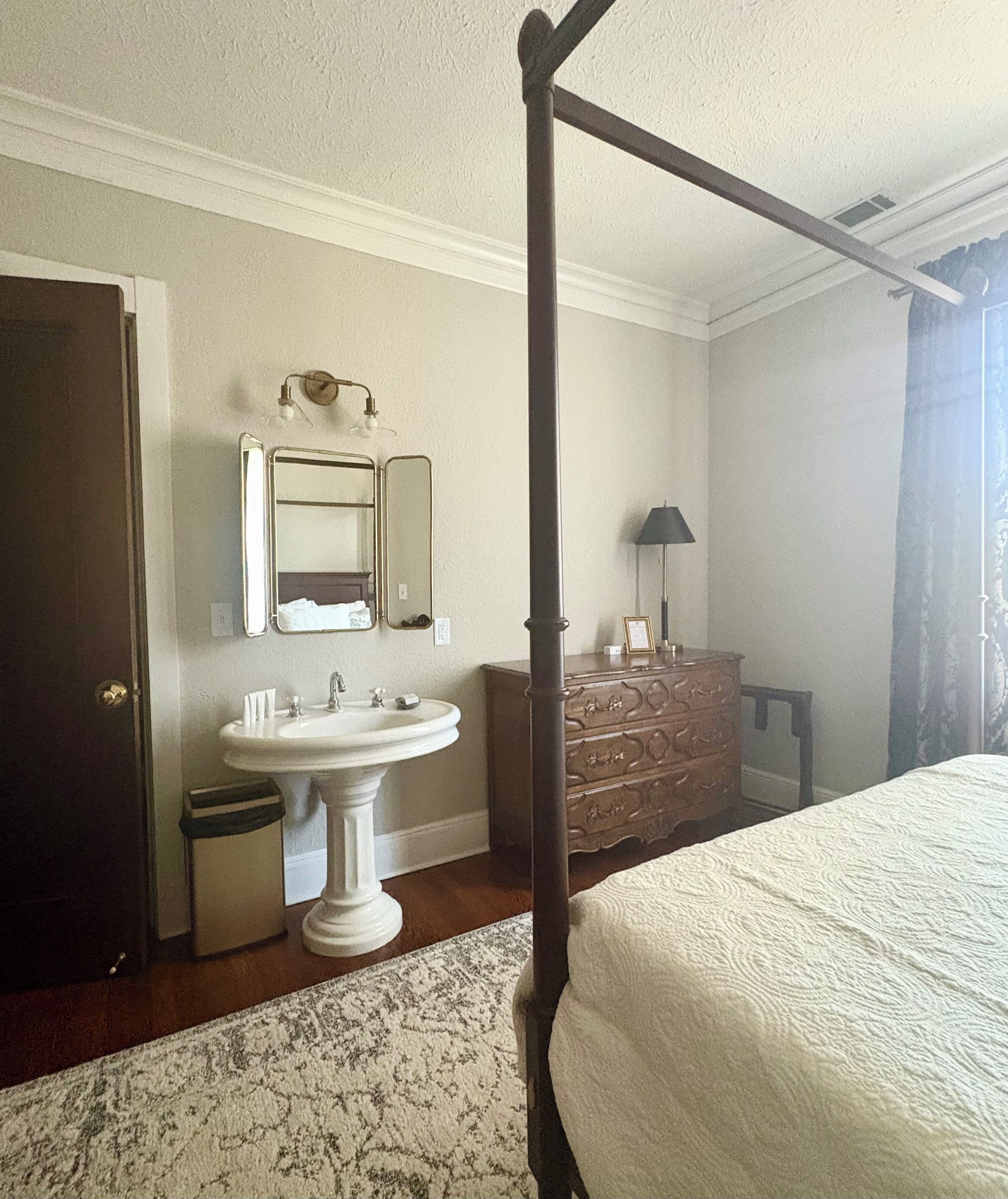 Un dormitorio con cama con dosel, un lavamanos pedestal, un aparador y un espejo.