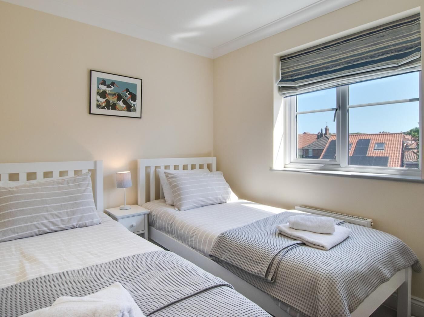 Une chambre d’hôtel avec deux lits, une fenêtre donnant sur des immeubles, une table de chevet avec une lampe et un tableau encadré au mur.