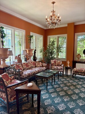 A room with a sofa, chairs, a coffee table, a chandelier, and large windows with plants.