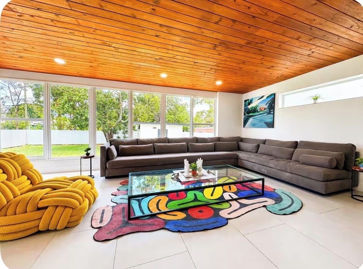 Een moderne woonkamer met een houten plafond, een hoekbank, een glazen salontafel en een kleurrijk vloerkleed.
