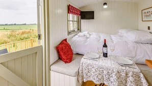 A bedroom with a bed, a table with a bottle and glasses, a red pillow, and a window with a view of the countryside. - 1 Bed in Market Rasen (oc-83846) (Faldingworth)