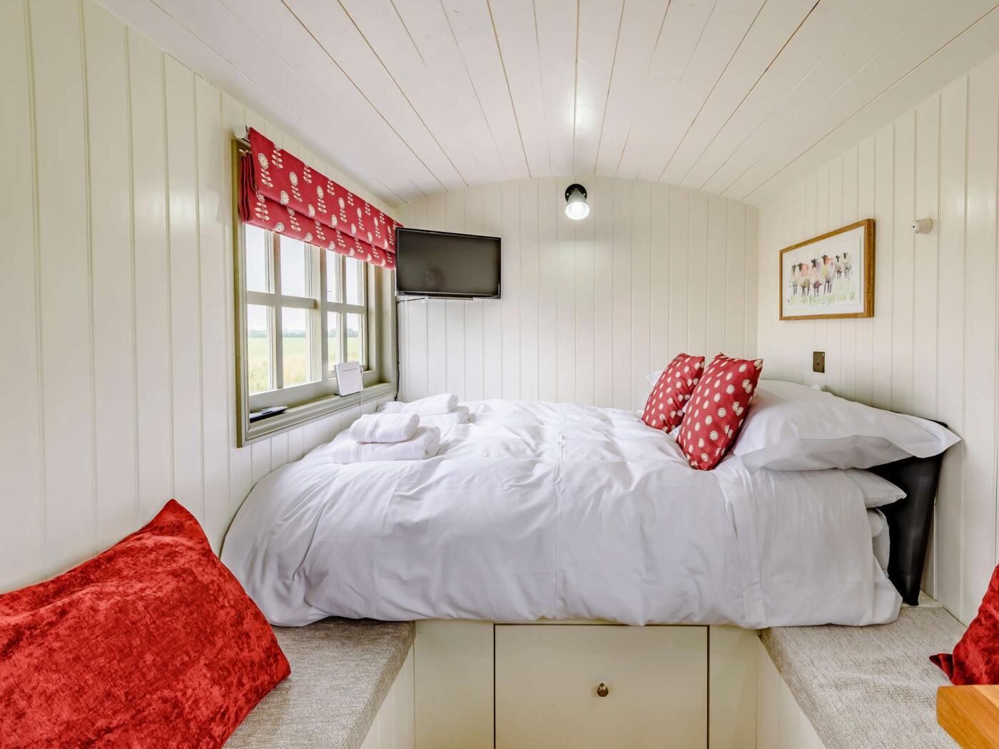 Une chambre à coucher avec un lit, une fenêtre ornée d’un rideau rouge, un téléviseur à écran plat fixé au mur et un tableau encadré accroché au mur.
