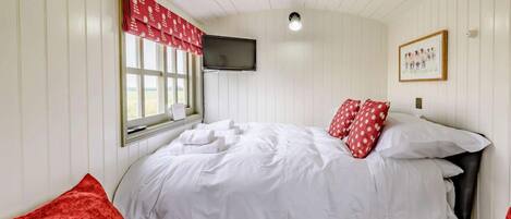 Une chambre à coucher avec un lit, une fenêtre ornée d’un rideau rouge, un téléviseur à écran plat fixé au mur et un tableau encadré accroché au mur.