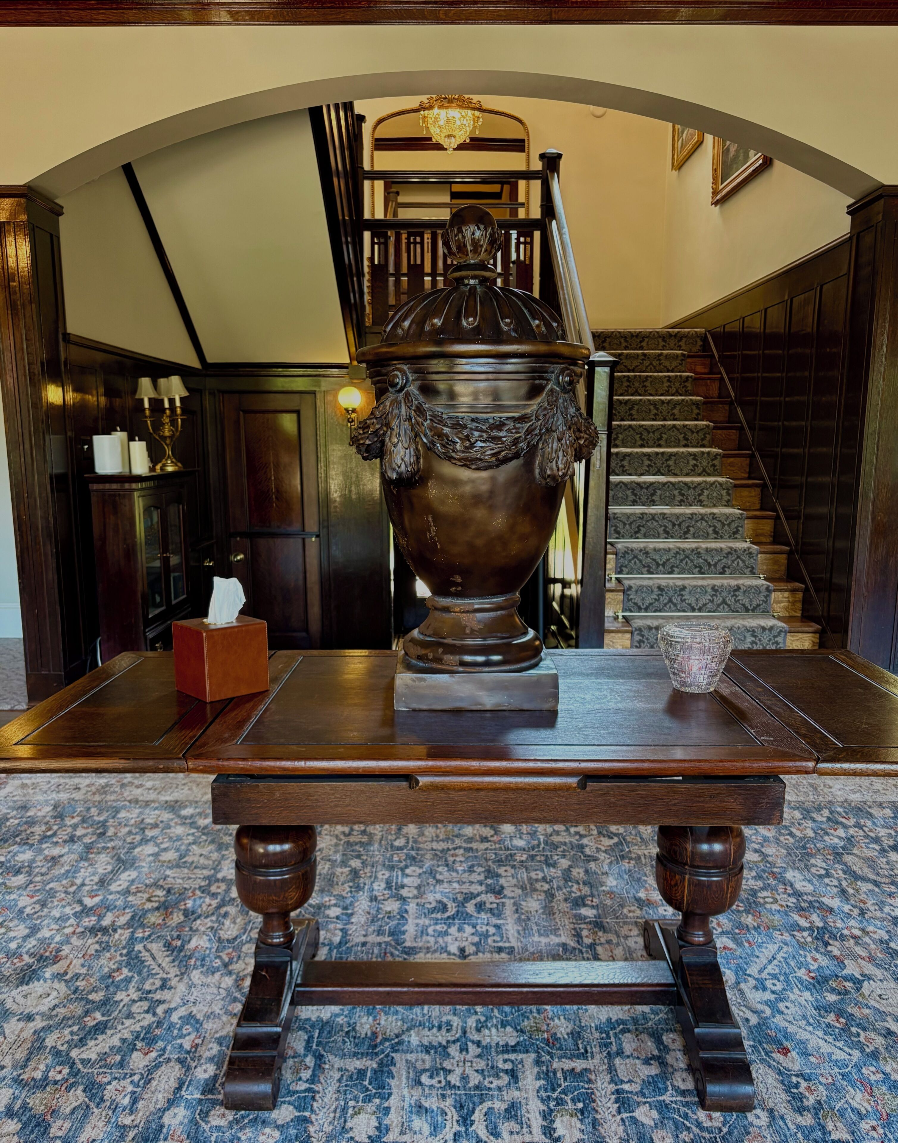 A grand room with a large ornate urn on a wooden table, a staircase with patterned carpet, and a chandelier.