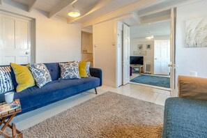 A living room with a blue sofa, a coffee table, and a rug. There is a door leading to another room with a television and a radiator. - 3 Bed in Polruan (oc-83821) (Fowey)
