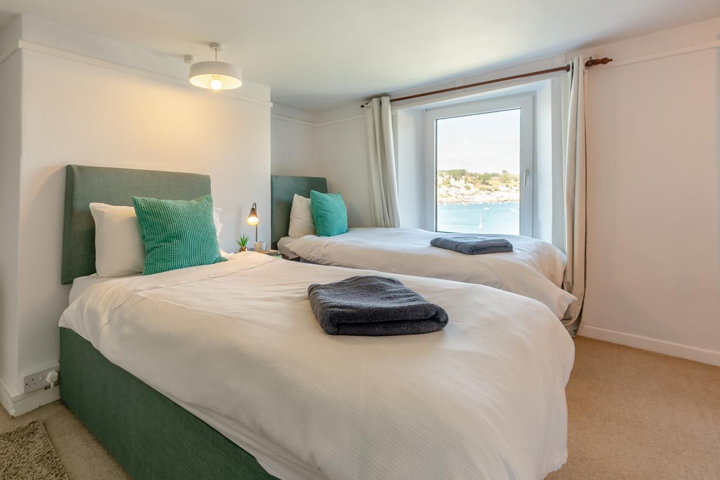 A hotel room with two beds, a window with curtains, a lamp, and a view of the sea.